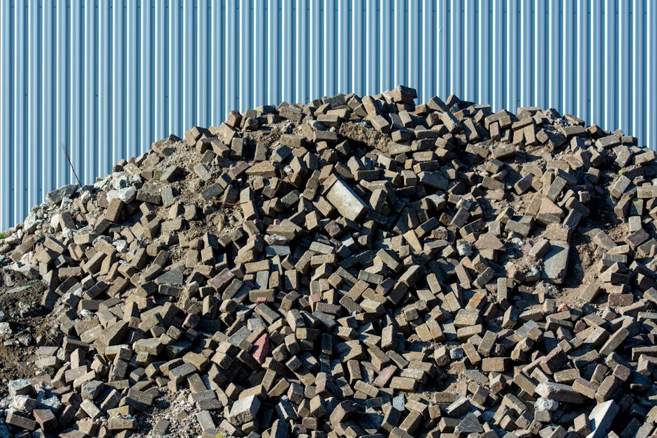 A large pile of irregularly shaped, dark grey to brown rectangular bricks and concrete debris is stacked against a corrugated metallic wall painted in light blue. The bricks have rough textures with some showing chipped edges and uneven surfaces, while a few pieces of concrete with exposed aggregate are visible among them. The heap sits directly on the ground, which appears to be a gravelly or dirt surface with some loose dust and smaller fragments scattered around the base. The metallic wall behind the pile extends vertically and reflects ambient light, creating subtle highlights along its ridges. The scene is brightly lit with natural sunlight, casting shadows that emphasize the jagged edges of the bricks. This arrangement of construction waste exemplifies separate, on-site waste materials that may be targeted by independent rubbish removal services like those offered by Rubbish Removal Pimlico, providing a visual representation of building debris that could be managed through private waste handling or alternative collection options rather than local authority disposal.