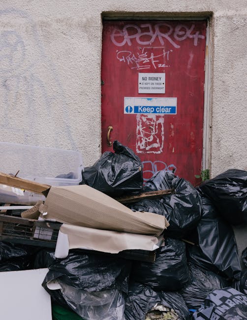 A red metal door with graffiti and stickers, including a white 'No Money' sign and a blue 'Keep Clear' notice, set into a beige concrete wall. In front of the door, there is a large pile of black rubbish bags filled with waste, some loosely tied and others partially open, with discarded cardboard and packaging materials stacked among them. The rubbish is situated on a paved surface, possibly a sidewalk or driveway, indicating a scene of illegal or unmanaged waste accumulation outside an urban building. The overall environment suggests an area where private waste disposal or rubbish removal services, such as those provided by Rubbish Removal Pimlico, might be required to handle such refuse responsibly, especially given the proximity to a building's exterior.