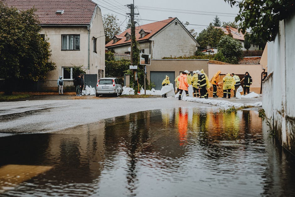 A group of workers wearing high-visibility yellow jackets, orange raincoats, and protective helmets are gathered on a residential street, engaged in clearing large white sandbags arranged along the sidewalk and roadway, likely as part of flood control or emergency waste management. The sandbags are stacked in several rows in front of clipped, older buildings with light-colored walls and tiled roofs, some with windows visible. A small silver van is parked nearby, partially blocking a driveway, with additional sandbags placed on the pavement beside it. The environment appears wet, with reflective surfaces on the road and a sizable pooled water area at the forefront, suggesting recent or ongoing flooding. In the background, trees and power lines are visible against an overcast sky. The scene is indicative of private or alternative rubbish handling associated with emergency waste disposal, aligning with independent collection efforts like those from Rubbish Removal Pimlico, servicing flood response cleanup and debris removal in the area.