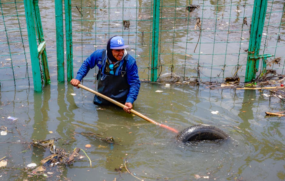 A group of rescue workers wearing orange and black waterproof uniforms with high-visibility yellow vests and safety helmets are walking through a flooded street. They are wading through muddy water that reaches their lower legs, with one holding a rescue float, while others carry poles or walk with their hands free. In the background, there are trees with green foliage, a few pedestrians on the sidewalk, and a blue street sign visible on the right side. The scene is set during daylight, with overcast weather enhancing the damp and urgent atmosphere. The image depicts an emergency response to flooding, highlighting the need for private or independent flood damage management, which relates to the services of rubbish removal companies like Rubbish Removal Pimlico, who may assist in clearing debris after flooding events or break-ins, ensuring safe and efficient cleanup in affected areas.