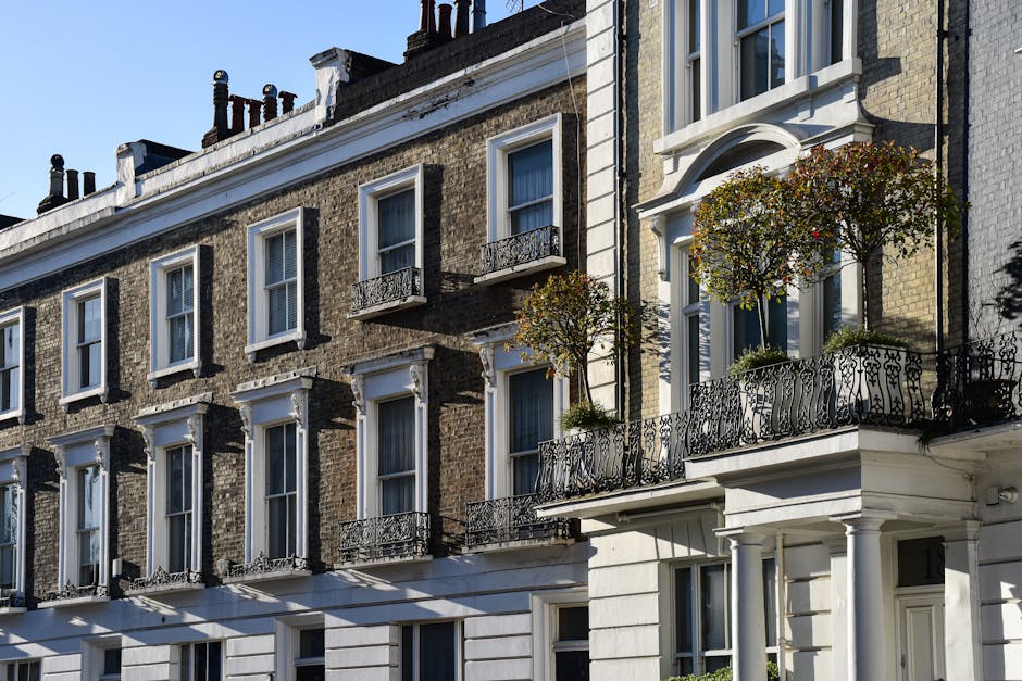 Photograph of a row of traditional Victorian-style residential buildings with multiple floors, featuring large sash windows with white frames. The upper floors display small, decorative wrought iron balconies from which potted plants and small trees are visible on the balconies on the right. The buildings are constructed with yellow brickwork, complemented by white architectural details around the windows and on the facade. The roofline includes chimney stacks and black-painted chimney pots. The natural sunlight highlights the textures of the brick and the ironwork, casting shadows on the building's facade. This scene reflects an urban environment with well-maintained exteriors, typical of city centre residential areas, and subtly aligns with the context of private property management or estate clearance services, as offered by Rubbish Removal Pimlico, in managing waste or clearance from such buildings.