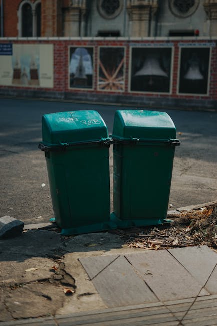 A collection of black trash bags filled with waste is piled in front of a red metal door set into a plain, beige concrete wall. The bags are stacked haphazardly, with some leaning on each other and others resting on discarded cardboard and packaging materials. The concrete wall shows signs of age and weathering, with small patches of discoloration and uneven surface texture. The red metal door features several graffiti tags and stickers, including a white sign indicating 'No Money' and a blue sticker instructing to 'Keep Clear.' The scene appears to be outdoors on a pavement or alleyway, suggesting an area where private rubbish collection or on-site clearance might take place, consistent with services such as those offered by Rubbish Removal Pimlico. The lighting is natural, highlighting the stark contrast between the dark bags and the muted tones of the surrounding environment, emphasizing the appearance of unmanaged waste collection typical of ad hoc rubbish disposal situations.