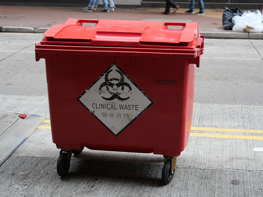 A large red plastic wheeled container designated for clinical waste, positioned on a concrete pavement outdoors. The container features a hinged lid with two handles on top and a white diamond-shaped biohazard symbol with black borders on its front, indicating the presence of hazardous medical materials. The label on the symbol reads 'CLINICAL WASTE' along with one line of text in Chinese characters beneath. The surface of the container has a slightly textured finish, typical of durable plastic, and the wheels at the base facilitate mobility across urban surfaces. In the background, a street scene is visible with pedestrians walking on a sidewalk opposite the container. To the right, there is a black rubbish bag and some discarded white plastic or paper material placed on the pavement. The setting appears to be an accessible public area where medical waste might be temporarily stored prior to specialized disposal, aligning with external clinical waste handling procedures and alternative waste management methods outside regular municipal rubbish collection.