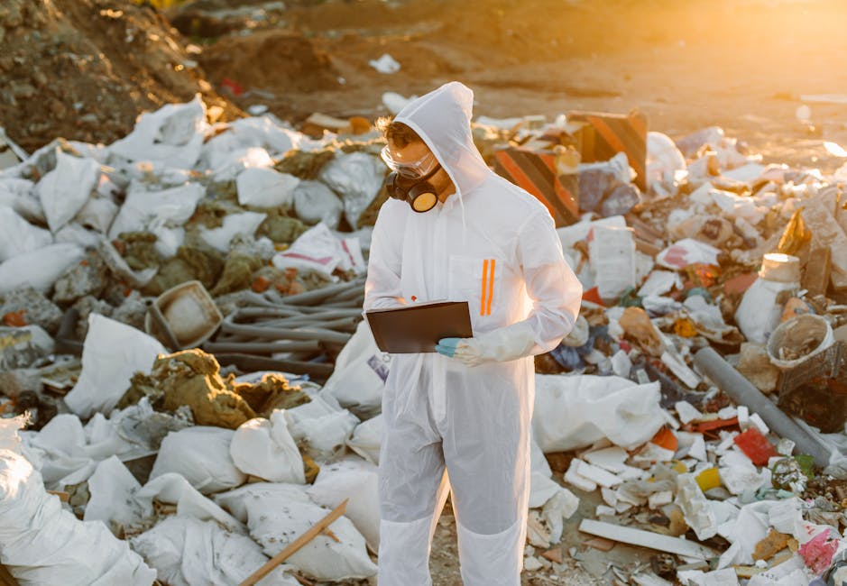 A person dressed in a white protective suit, gloves, goggles, and a face mask is standing amidst a large piles of mixed waste and debris, including broken insulation, plastic fragments, paper, and discarded building materials, on an outdoor site during the daytime with warm sunlight. The individual is holding a clipboard and appears to be inspecting or recording details related to waste management or hazardous waste disposal. The environment suggests a contaminated or industrial area with scattered rubbish, and the scene emphasizes the importance of professional waste handling and proper disposal processes that Rubbish Removal Pimlico may facilitate as part of alternative rubbish collection services. The image highlights a controlled approach to managing significant waste accumulation in non-residential or site clearance contexts, conveying the necessity of safety equipment and meticulous documentation in waste removal operations involving potentially hazardous materials.