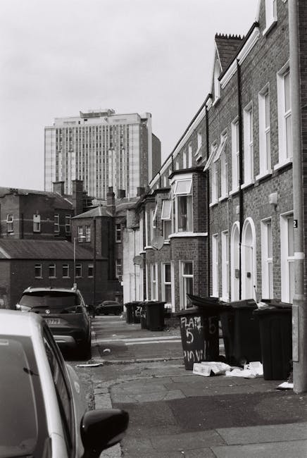 A row of terraced brick houses on a residential street, with front doors and small bay windows facing the pavement. Several black wheelie bins and recycling containers are placed along the edge of the pavement, some with lids open or slightly ajar. In the foreground, part of a parked vehicle is visible on the left side of the image. The street appears tidy, with minimal clutter aside from the bins. In the background, there is a large multi-storey modern office or apartment building made of glass and concrete, contrasting with the older residential buildings in the foreground. The scene is depicted in grayscale, with diffuse daylight and an overcast sky, providing an observational view of typical private waste collection arrangements non-sponsored by local authorities. The setting and visible waste bins suggest an environment where independent rubbish collection services may be used, such as Rubbish Removal Pimlico, for handling refuse outside scheduled municipal collections.