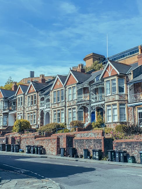 A row of terraced brick houses on a residential street, with front doors and small bay windows facing the pavement. Several black wheelie bins and recycling containers are placed along the edge of the pavement, some with lids open or slightly ajar. In the foreground, part of a parked vehicle is visible on the left side of the image. The street appears tidy, with minimal clutter aside from the bins. In the background, there is a large multi-storey modern office or apartment building made of glass and concrete, contrasting with the older residential buildings in the foreground. The scene is depicted in grayscale, with diffuse daylight and an overcast sky, providing an observational view of typical private waste collection arrangements non-sponsored by local authorities. The setting and visible waste bins suggest an environment where independent rubbish collection services may be used, such as Rubbish Removal Pimlico, for handling refuse outside scheduled municipal collections.