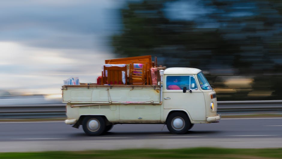 A vintage cream-colored van is seen traveling along a road with rapid motion, creating a blurred background of trees and sky that emphasizes its movement. The van's exterior appears slightly weathered, with visible signs of age on its paintwork and metal surface. Loaded onto the flatbed area at the back are various household furniture items, including a wooden dining chair, a small table, and a cardboard box, all secured in place. The furniture exhibits different finishes, with some pieces showing polished wood and others more matte textures, reflecting different material types. The items are stacked and arranged to maximize space, with the wooden chair and table leaning slightly to the side and the box positioned nearby. The environment suggests an outdoor setting, possibly on an urban or suburban street, with the motion blur indicating a high-speed transit. This scene illustrates private waste transportation, characteristic of independent rubbish removal services, where household items are loaded for disposal or recycling, consistent with the services provided by Rubbish Removal Pimlico.