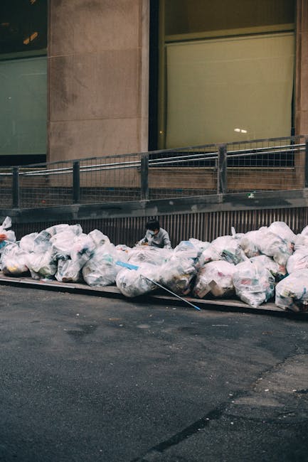 A row of large, transparent plastic rubbish bags filled with mixed waste, placed along the edge of a paved city street outside a modern building. The bags, appearing somewhat disorganized and bulging with varied contents such as paper, plastic, and other refuse, are situated adjacent to a metal railing and a section of the building's concrete wall. Two individuals are visible behind the bags, partially obscured by the refuse, appearing to be sorting or managing the waste. The background features large windows with dark frames and reflective glass, and the scene is lit with ambient natural light, indicating an outdoor urban environment. This setup suggests a private waste disposal or alternative rubbish collection near commercial or office premises, in alignment with independent rubbish removal services often employed for on-site clearance or non-local authority waste handling, as offered by services like Rubbish Removal Pimlico.