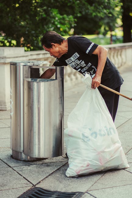 A middle-aged man with dark hair, dressed in a black T-shirt with white text and dark pants, is seen bending over a stainless steel trash bin on a paved sidewalk next to a low stone wall. He is holding a large white rubbish sack labeled with a faint logo or text, and appears to be discarding waste into the bin. The scene is set outdoors in a lush area with green trees and shrubbery in the background, and the lighting suggests daytime with natural sunlight. The man is using a tool, possibly a broom or stick, to assist with rubbish collection, indicating an active waste clearance effort. This image exemplifies private rubbish disposal and on-site waste handling services, often provided by companies like Rubbish Removal Pimlico, avoiding reliance on local authority collection.