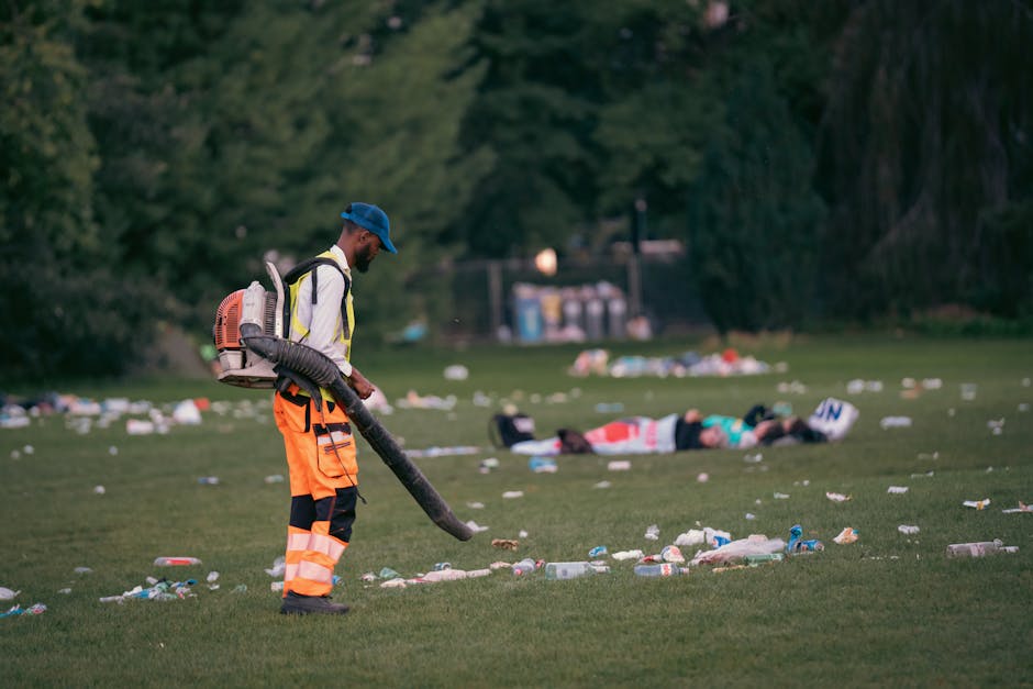A waste management worker dressed in a high-visibility orange and black uniform with reflective strips, wearing a blue cap, is using a leaf blower to clear litter from a grassy park area. The worker is positioned in the foreground, slightly to the left, with their head tilted downward towards the ground. Behind them, scattered trash including plastic bottles, paper, and other debris is visible across the lawn. In the background, there are two individuals lying on the grass, with trees and a small structure with recycling bins and waste containers further back, indicating an outdoor public space. The scene appears to be set during the late afternoon or early evening with diffuse natural lighting, and the environment suggests an area that may require routine rubbish removal or cleaning, supported by the presence of the rubbish removal company, Rubbish Removal Pimlico, specializing in on-site waste clearance services.