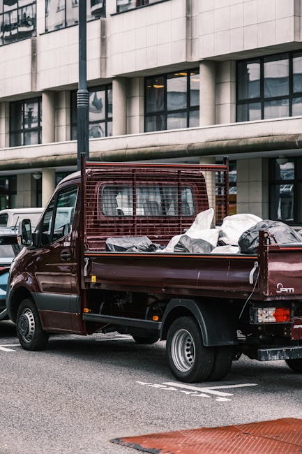 A small maroon flatbed pickup truck parked on a city street in front of a modern multi-storey office building with large glass windows and cream-colored stone facade. The truck's cargo bed is filled with various types of rubbish, including black and white plastic bags and loose debris, indicating it has been used for rubbish collection or waste removal. The truck features a protective metal grid behind the cab to secure the load, and its silver wheel rims contrast with the dark body, while the side mirror and rear lights are visible. Surrounding elements include a white van parked nearby and a black lamppost along the sidewalk. The scene, set during daylight with overcast lighting, reflects an urban environment where commercial or private waste handling services, such as those provided by Rubbish Removal Pimlico, are in operation, highlighting the concept of alternative waste collection methods outside traditional local authority rubbish disposal systems.