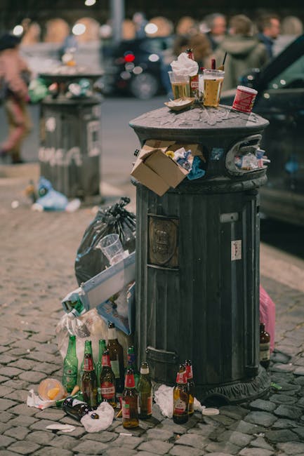 A black cast-iron public waste bin positioned on a cobblestone sidewalk, with its lid open and overflowing with waste. On top of the bin, there are used paper coffee cups, some with plastic lids, and crumpled paper napkins. Attached to the side of the bin is a cardboard box filled with discarded rubbish. Surrounding the base of the bin, there are numerous empty glass beer bottles, both green and brown, some with caps still on, and scattered food wrappers. In the background, a blurred city street scene includes a parked car on the right, a grey trash bag leaning against the bin, and a recycling or waste collection site in the distance, indicating an urban environment. The overall scene captures littering near a busy street, emphasizing the importance of proper rubbish handling and disposal as promoted by waste management services.